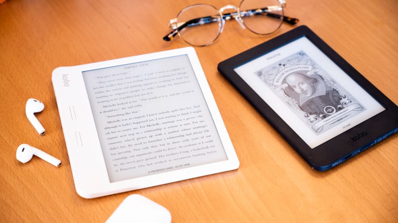 Two Kobo e-readers sit on a desktop with a pair of glasses and a set of AirPods.
