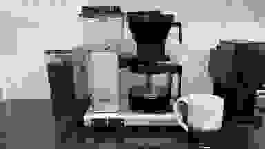 The Technivorm Moccamaster coffee pot full of coffee sitting on a black counter with a container of beans to the left, a coffee grinder to the right, and a mug in front that says "Coming in hot."