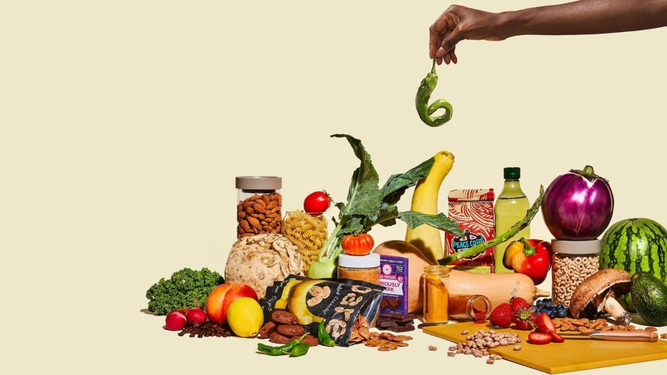 A hand holds a curly green pepper above a pile of sustainable groceries including misshapen produce and eco-friendly packaged goods.