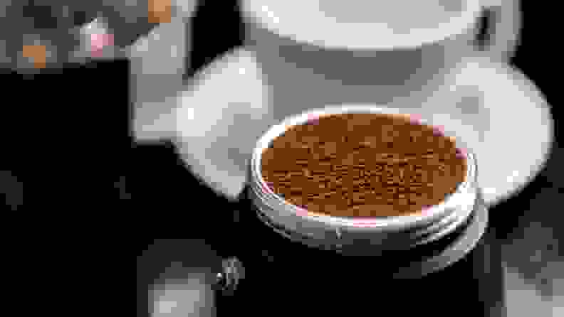 A filter filled with coffee grounds sits in the bottom compartment of a moka pot in the foreground, with a white coffee cup and saucer in the background.