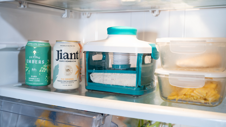 A Noya Tofu Press sitting on a fridge shelf, alongside cans of kombucha and tupperware containers of leftovers.