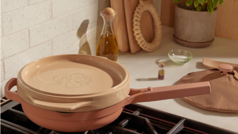 A pink plate is nestled on top of the Always cooking pan on the stove; and the pan lid is wrapped with cotton cloth.