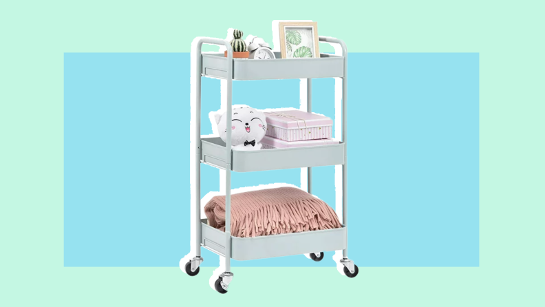 White 3-tier utility cart loaded with blankets, potted cactus, picture frame, stuffed animal and alarm clock.