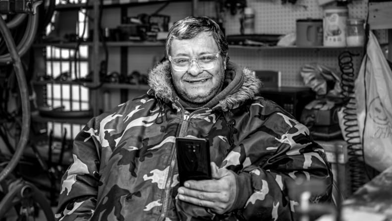 A man in a camouflage jacket sits in his bicycle repair shop, browsing on his smartphone.