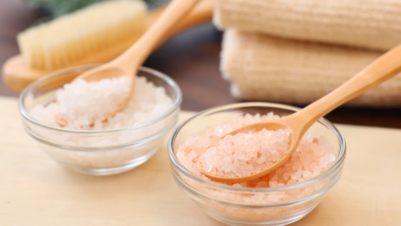 Two bowls with bath salts with wooden spoons next to beige towels and a dry brush