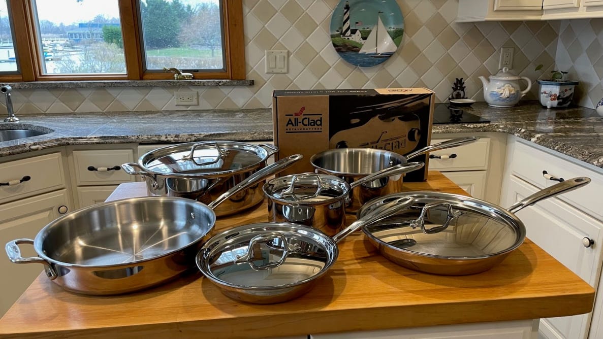 All Clad's D3 Stainless Steel Cookware set sitting on top of wooden island in the middle of modern kitchen.