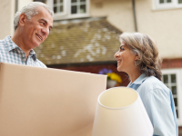 Two people moving into a house stand outside carrying a box and a lamp.
