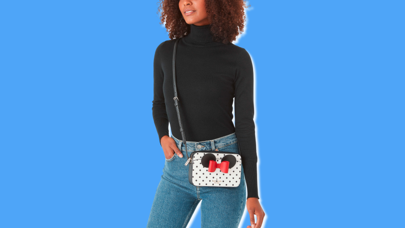 A woman wearing a black turtleneck wearing a small crossbody bag with black polka dots on a while background with a red bow and black Minnie ears.