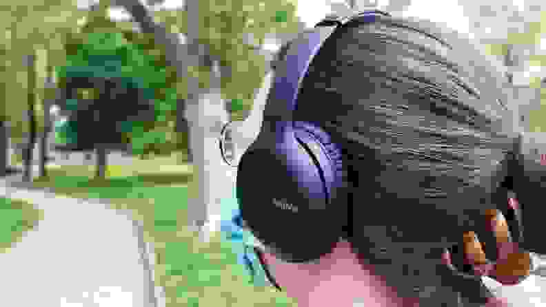 A person wearing a pair of Sony's WH-CH710N headphones with their hair in a bun and clipped, standing in a park near a walking path.