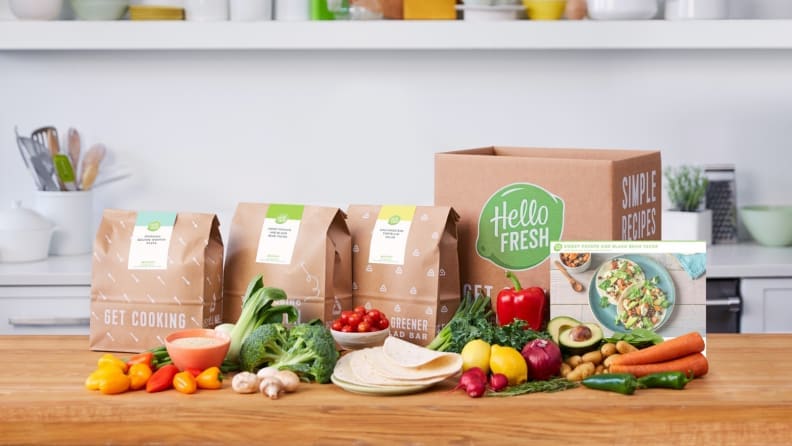 A HelloFresh meal kit box on a wooden kitchen counter surrounded by fresh produce and paper meal kit bags.