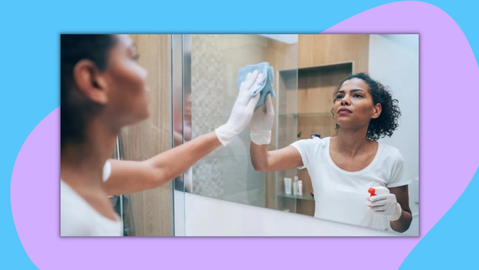 Woman wearing clear gloves and using spray bottle and cloth to wipe off mirror.