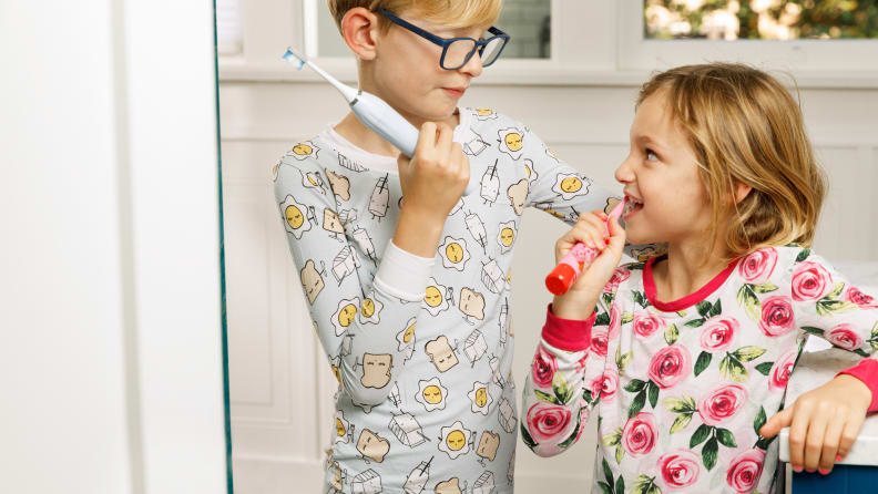 A boy and a girl wear pajamas and smile at one another while holding toothbrushes.