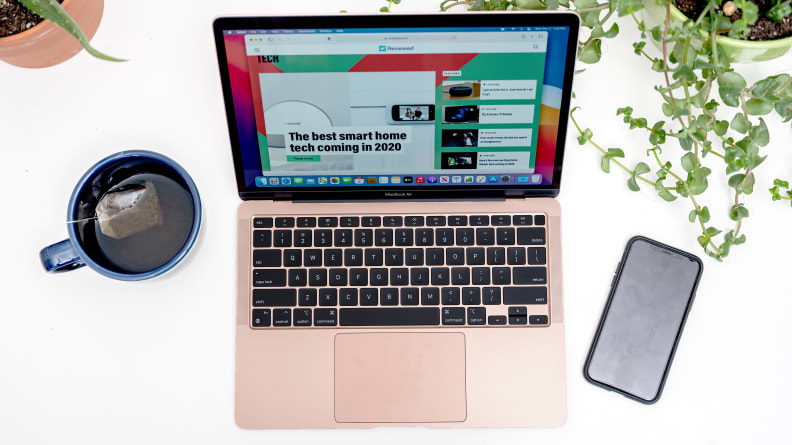 A MacBook Air is flanked by a cup of tea and a smartphone. The laptop is open to the Reviewed website.