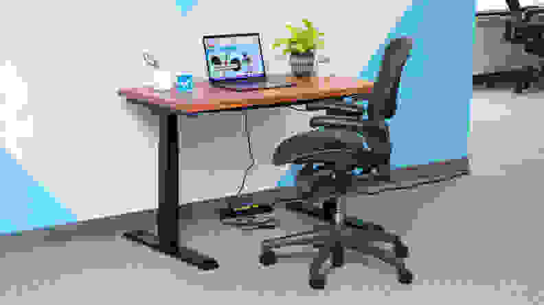 The Flexispot E7 standing desk  in front of a blue and white wall with an office chair in front of it and a laptop on the desk.