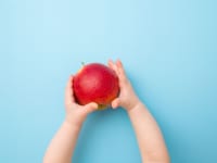 A toddlers hands extended holding a red apple against a light blue background.