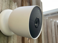 Google's Nest Cam (battery) home security camera hangs on a wood fence outdoors.