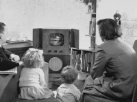 A family of four - a mother, father, and two small children - sit around a small black-and-white television screen in the 1950s.