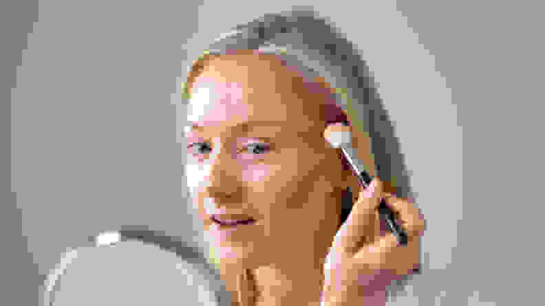 A middle-aged person looks into a round mirror only wearing foundation on their skin while holding a makeup brush to their face.