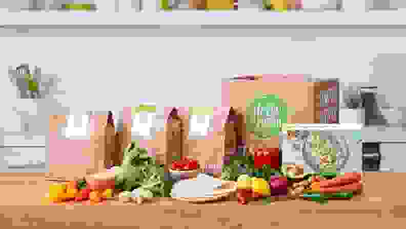 A HelloFresh meal kit box on a wooden kitchen counter surrounded by fresh produce and paper meal kit bags.