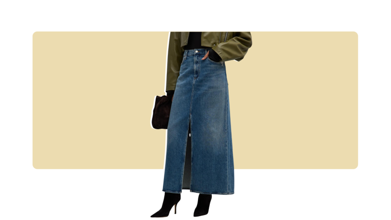 A model seen from the waist down wearing an ankle-length blue denim skirt with a front slit, paired with black boots, an olive jacket, and a bag.