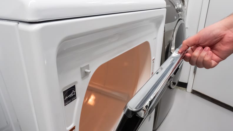 A close-up of a hand reaching into frame to open the Maytag MED7230HW dryer's hamper door, which swings from top to bottom.