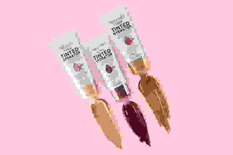 Three Wet n Wild Bare Focus Tinted Hydrators in light, medium, and dark skin tones lay on a light pink background with swatches of the formula coming out of the tubes.