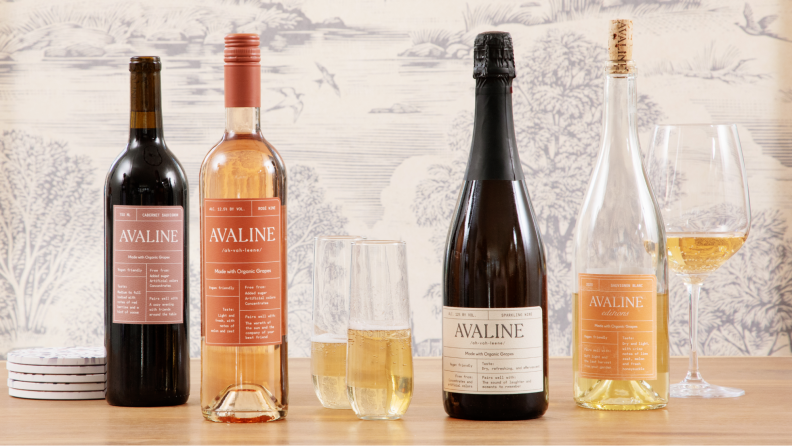 Four bottles of Avaline with glasses of wine and a few glasses filled with white whine and sparkling wine sit on a wood table.
