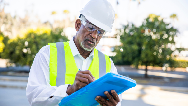 Technician wearing vest and hard hat outdoors writing on clipboard.