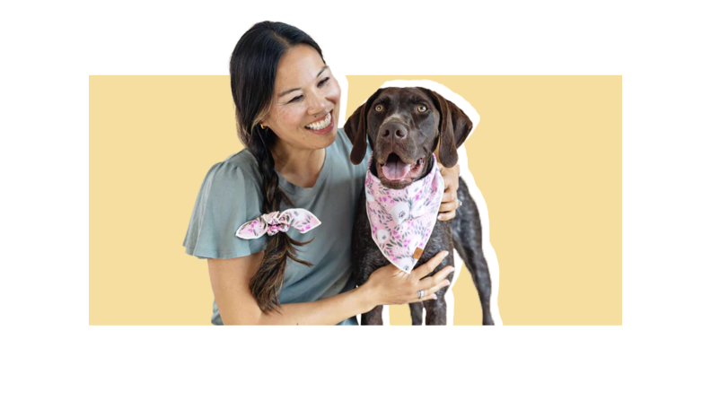 A woman wearing a pink patterned scrunchie tied around her braid next to a chocolate lab wearing a bandana of the same pattern, both against a light gold backgroudn.