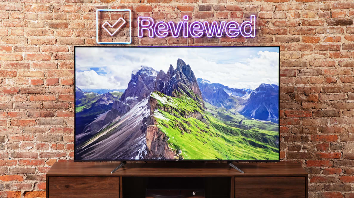TCL 65Q650G LED TV with green landscape with mountains on screen indoors in front of brick wall.