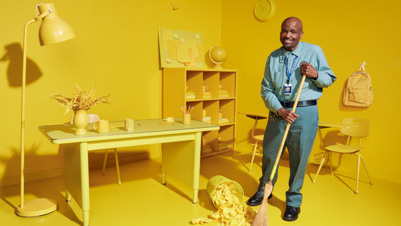 A photograph of actor William Stanford Davis dressed as Mr. Johnson, the custodian from Abbott Elementary, sweeping up a yellow room.