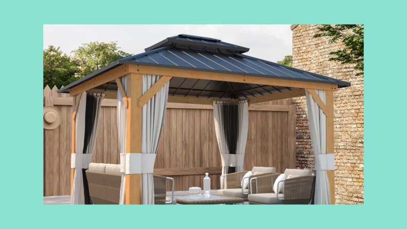 A wood gazebo with a black roof and curtains outdoors in front of a fence