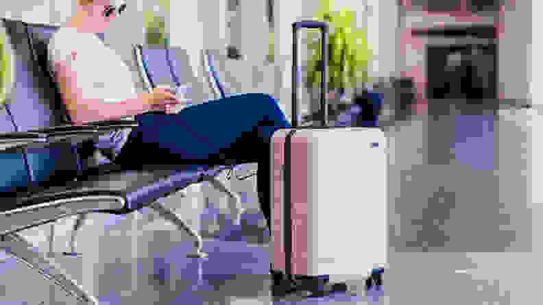 A woman sitting at an airport with a pink hard sided rolling suitcase next to her