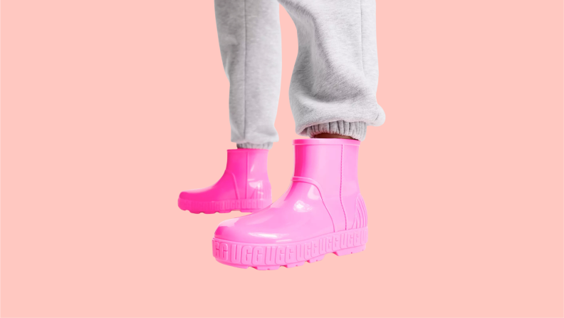 A close up of a person wearing a pair of bright pink Ugg rainboots featuring the Ugg logo on repeat around the midsole of the shoe.