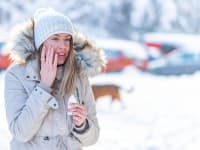 A person wearing a winter jacket and hat in a snowy location holds a tube of suncreen and touches her hand to her cheek.