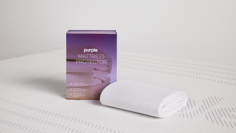 The Purple Mattress Protector in white rolled up next to its purple packaging on a white mattress.