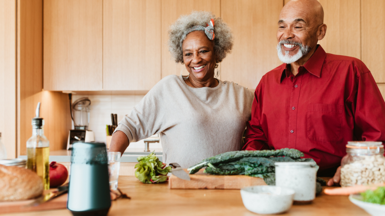 A couple speaks to a smart speaker as they make a meal in their kitchen.