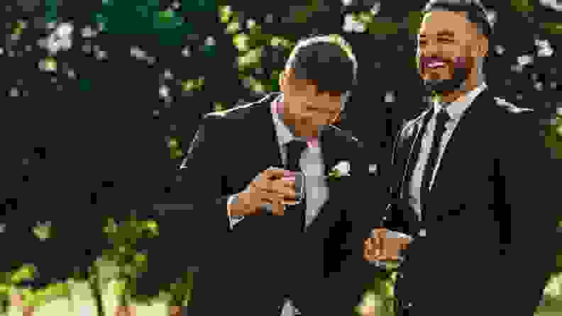 A groom and his best man dressed in black tuxedos hold cocktails and laugh