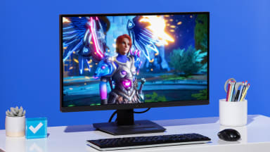 The Asus ROG Strix XG27ACS, a black gaming monitor, on a white desk against a blue wall