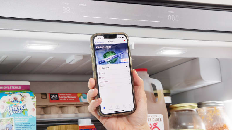 A hand holds a phone and shows the Bosch app in front of a Bosch refrigerator