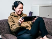 Person seated on couch holding portable gaming device and smiling at screen.