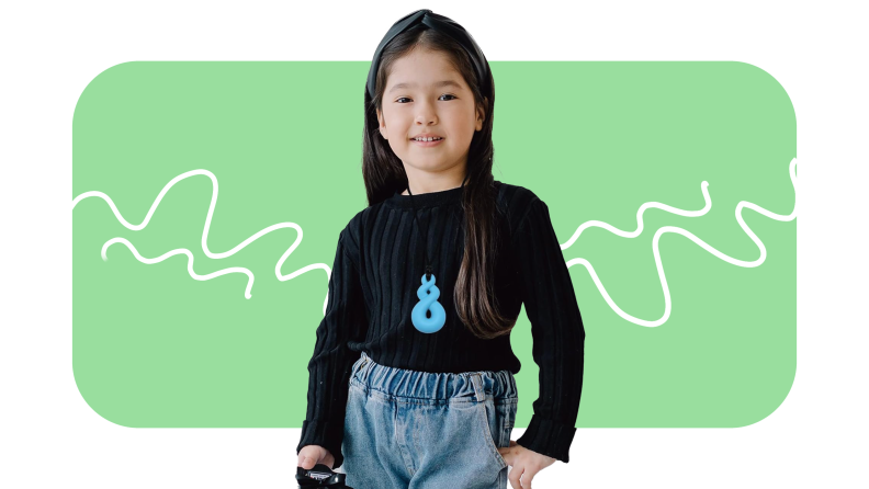Small child poses while wearing blue chew necklace around neck, blue jeans and black sweater.