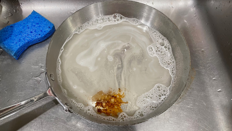 A stainless steel skillet filled with soapy water inside of a sink next to a blue sponge.