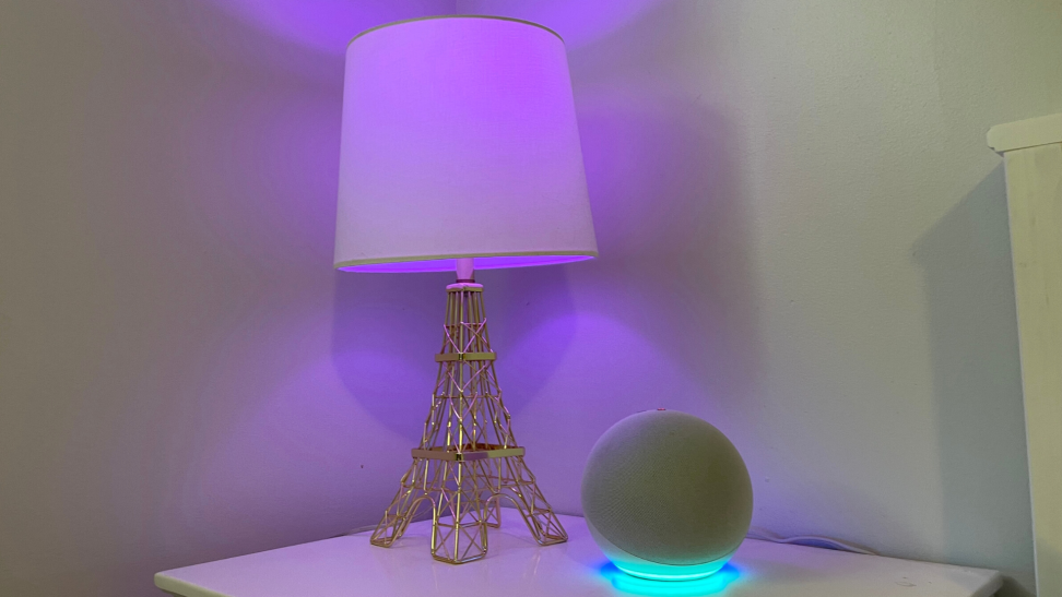 Amazon's Echo Dot (4th Gen) sits on a white nightstand next to a lamp with a purple smart bulb.