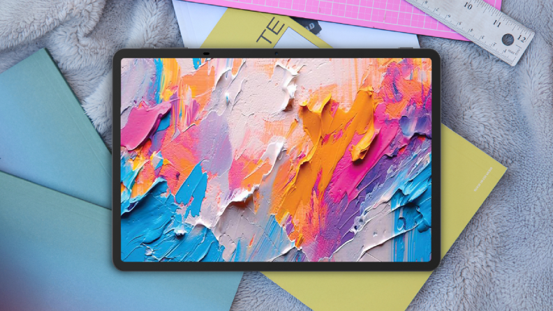 A sleek tablet displaying a vibrant abstract painting with bold, colorful brushstrokes, placed on top of soft textured fabric and surrounded by notebooks and stationery.