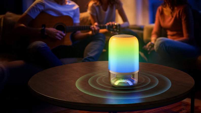 A vibrant smart speaker with a glowing rainbow light display sits on a wooden table, emitting sound waves, while a group of friends enjoys an evening, with one person playing guitar in the background.
