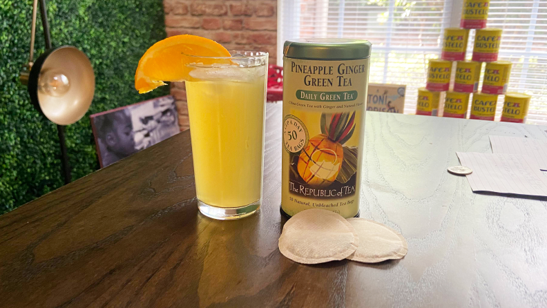 A canister of Republic of Tea Ginger Green Tea sits on a table next to a glass of the beverage