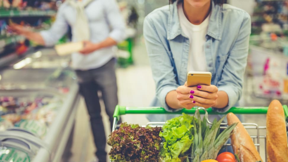 Girl leaning on shopping cart, using a mobile phone and smiling, in the background a man is choosing food.