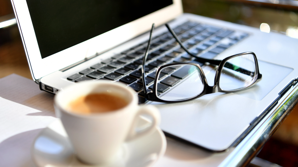 A pair of eyeglasses rest on the keyboard of a laptop next to a hot drink.