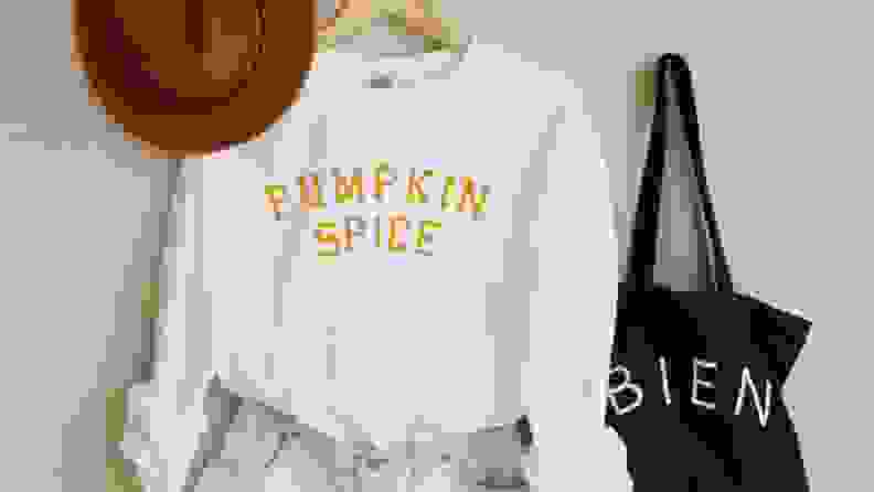 A white sweater that says 'Pumpkin Spice' next to a hat, tote bag, and jeans